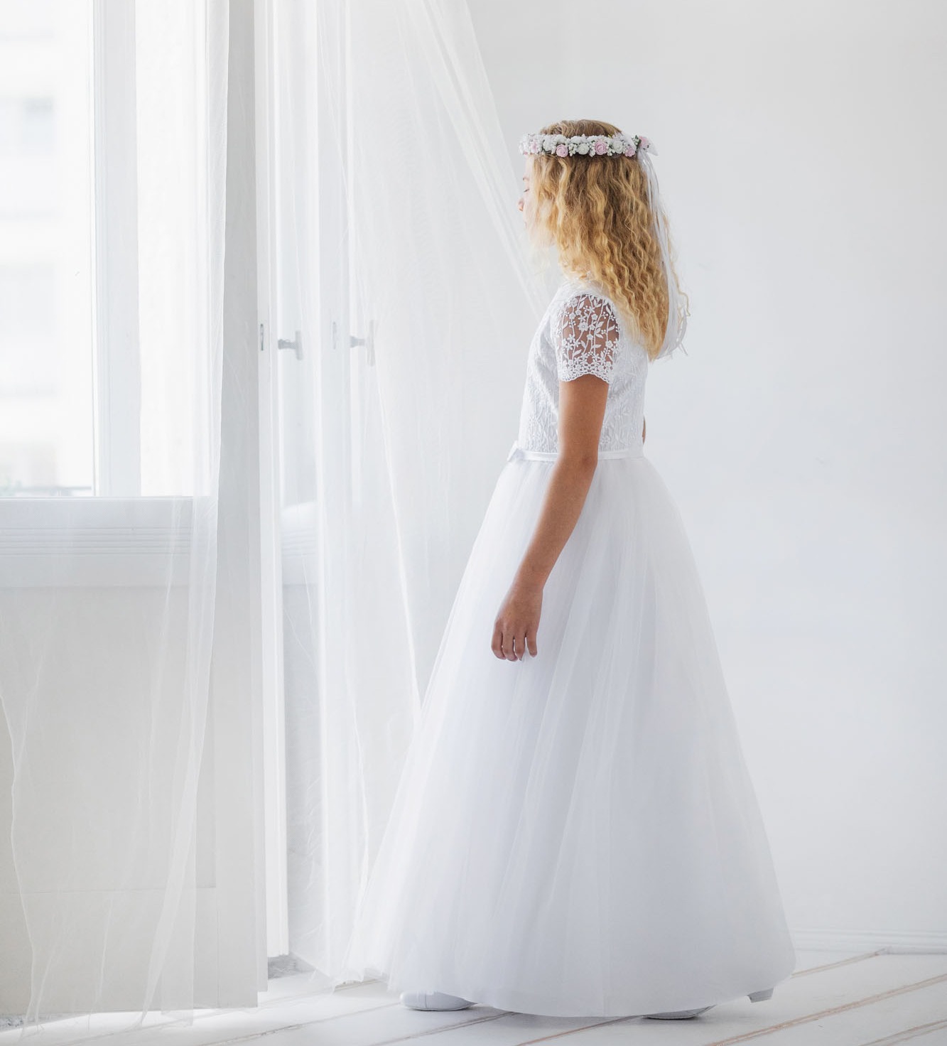 Dziewczynka w sukience komunijnej z tiulem i koronką i we wianku komunijnym z różowymi kwiatami